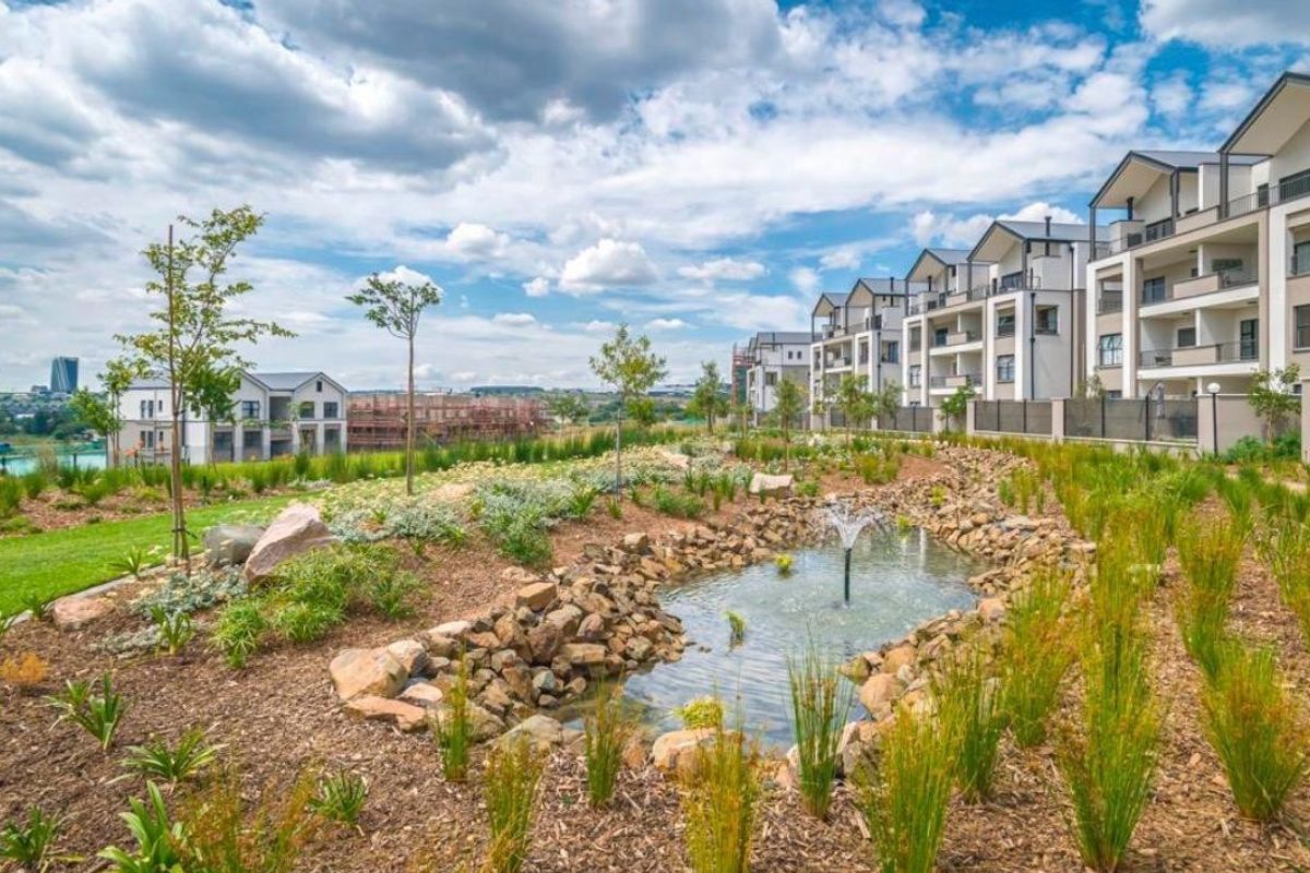A well-maintained communal garden area featuring a rock-lined water fountain, surrounded by modern multi-story apartment buildings with private balconies.