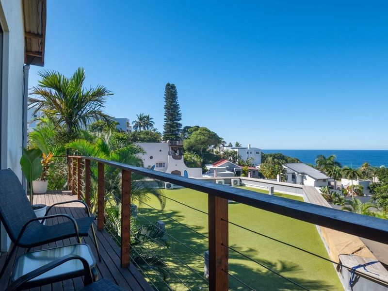 A spacious balcony featuring timber decking and a modern wire balustrade, offering elevated views of the surrounding neighbourhood and the ocean.