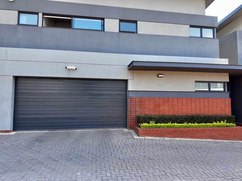 Modern townhouse exterior featuring a sleek automated garage door, paved driveway, and a decorative brick planter box.