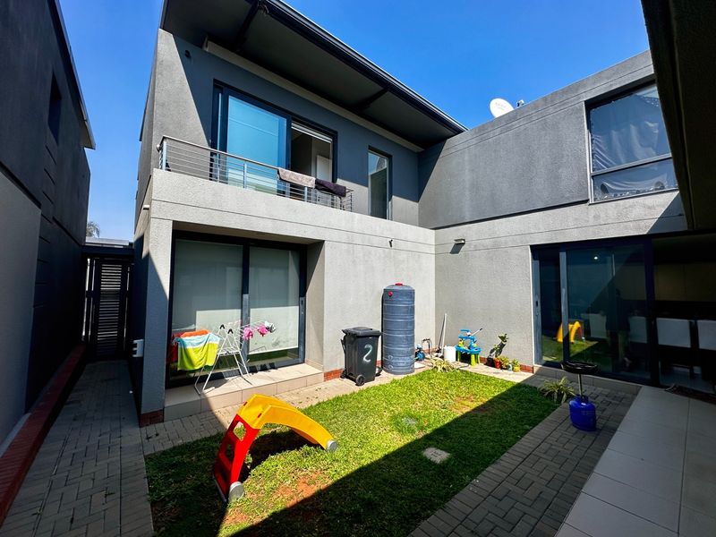 A modern townhouse rear courtyard featuring a paved patio area, small lawn, and an upper-level balcony with sliding door access.