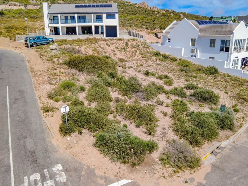 An aerial view of a vacant residential plot situated on a corner, featuring natural vegetation and direct street access in a developed neighborhood.