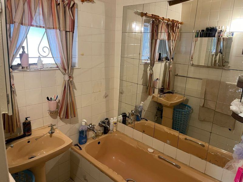 A dated bathroom featuring a pedestal washbasin, built-in bathtub, and wall-to-wall mirror, with original ceramic tiling throughout.