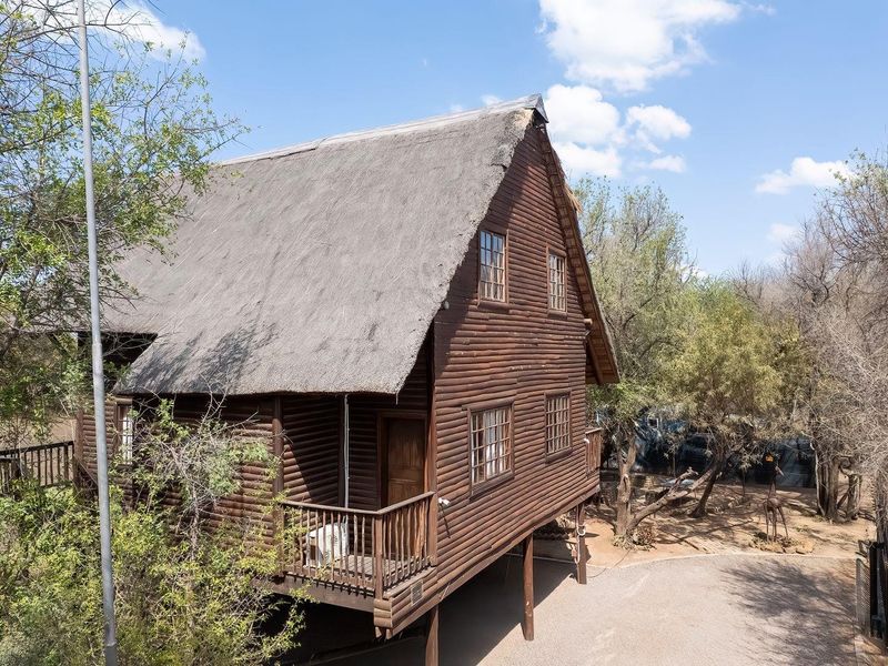 A rustic stilted log cabin featuring a traditional thatched roof and a wooden balcony, set within a natural bushveld environment.