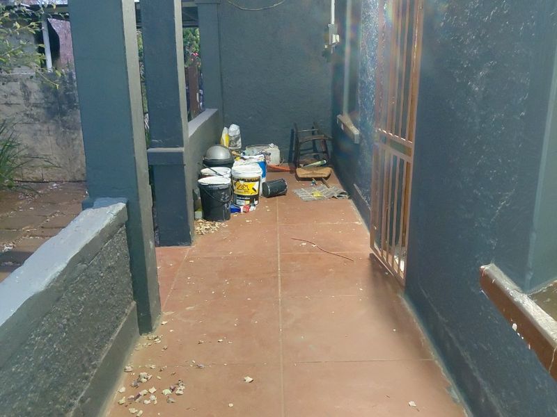 A covered outdoor patio area featuring tiled flooring, painted masonry columns, and a security gate, currently requiring maintenance and cleaning.