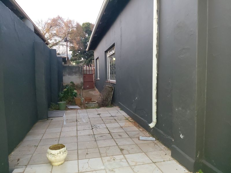 A side exterior area featuring a tiled patio, dark painted walls, and a functional downpipe, leading towards a rear security gate.