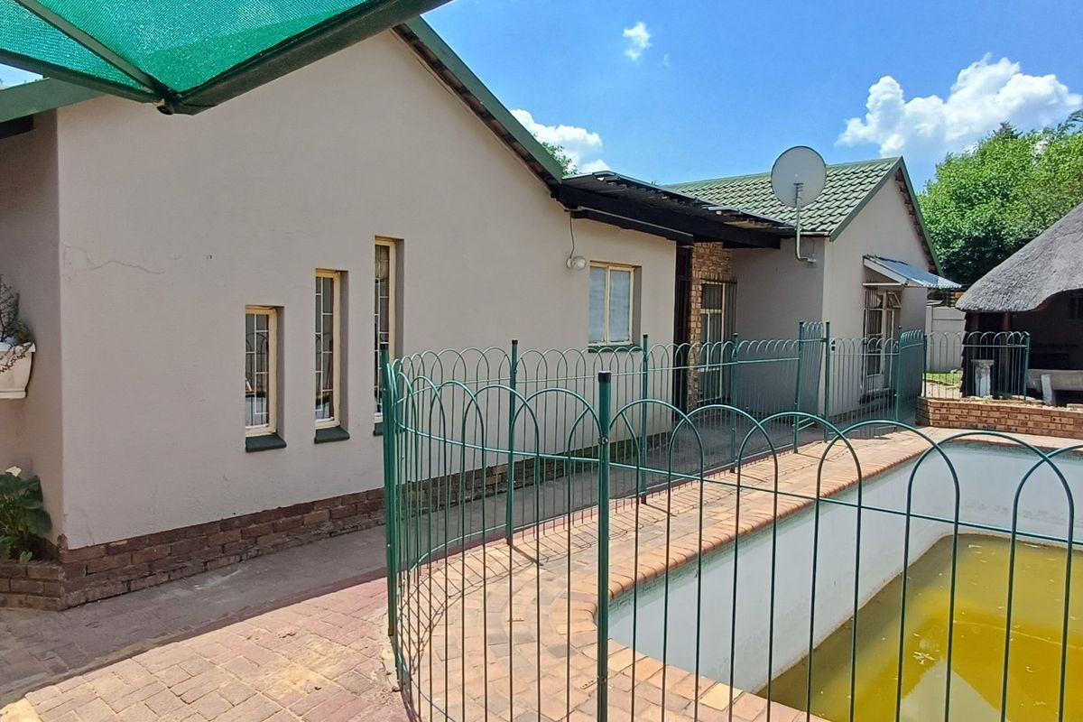 Rear exterior view featuring a swimming pool with safety fencing, paved patio area, and a traditional thatched lapa structure.