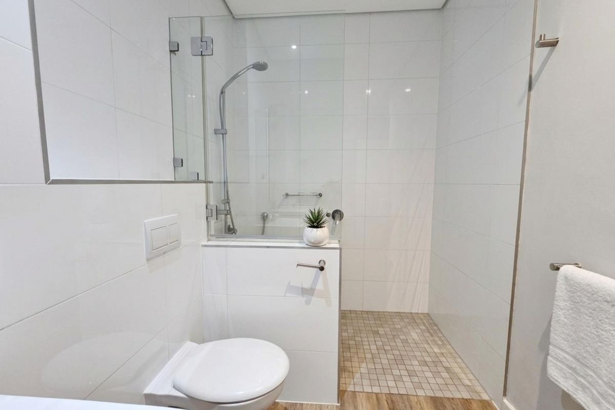 Modern bathroom featuring a wall-mounted toilet, a glass-enclosed shower with tiled flooring, and clean white wall tiling.