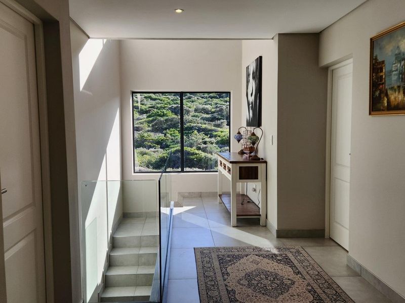 A bright, modern hallway featuring tiled flooring, a glass balustrade staircase, and a large window offering scenic natural views.