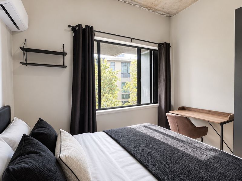 A modern bedroom featuring an exposed concrete ceiling, air conditioning unit, and a built-in wardrobe with a view of the surrounding neighborhood.