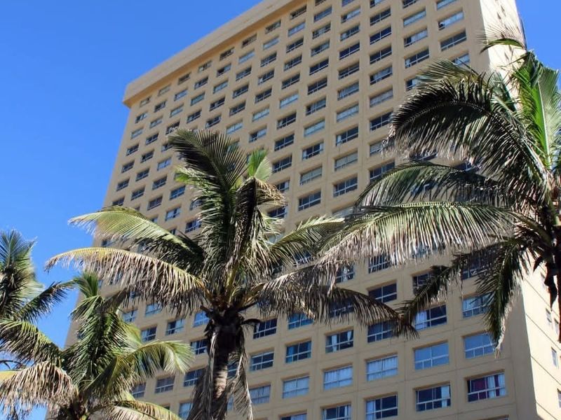 A low-angle view of a multi-story high-rise apartment building facade featuring a uniform grid of windows and palm trees in the foreground.