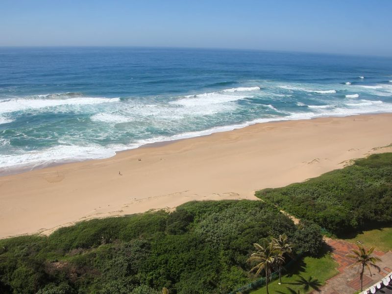 A high-angle panoramic view of the coastline, featuring sandy beach, ocean waves, and coastal vegetation.