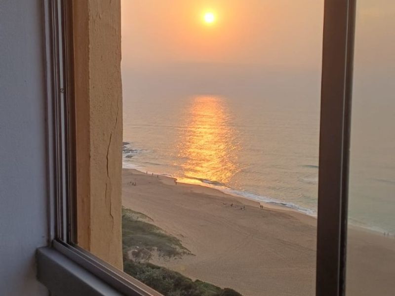 A scenic view of the ocean and beach at sunset, framed by a sliding window.