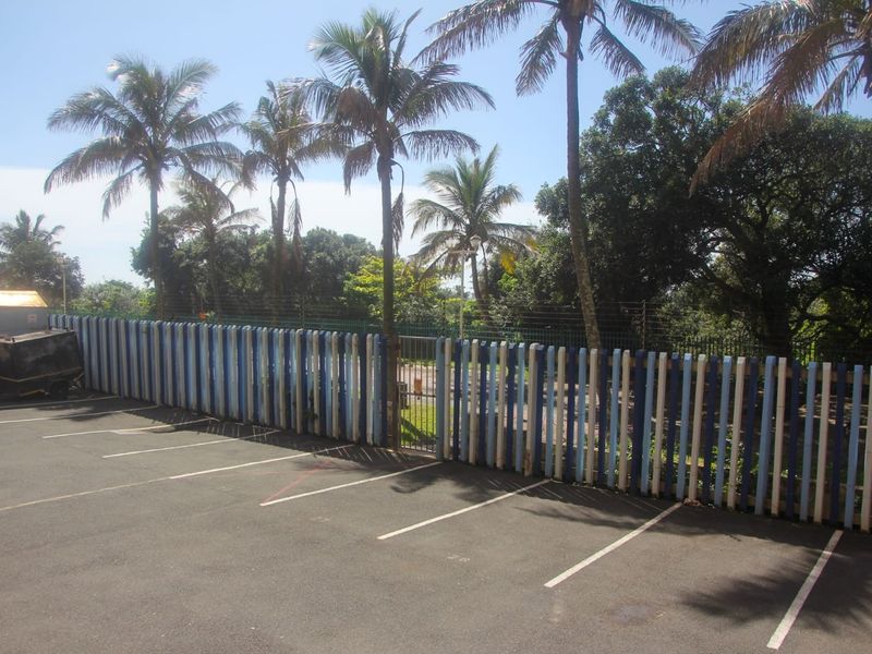 Paved parking area featuring a painted steel palisade fence with integrated security gate and electric fencing, surrounded by mature palm trees.