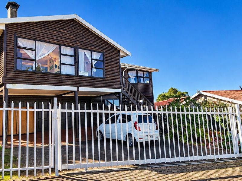 A two-story timber-clad house featuring a metal picket fence, paved driveway, and an external staircase, presented in a clear, sunny exterior view.
