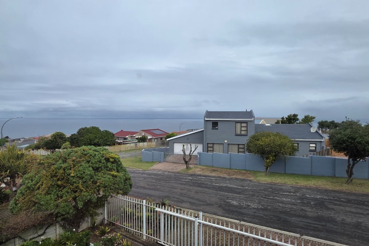 Elevated view of a residential property featuring a grey exterior, paved driveway, double garage, and perimeter wall with a distant ocean horizon.