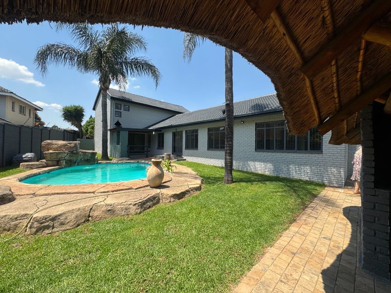 A well-maintained backyard featuring a swimming pool with a natural rock water feature, surrounded by a lawn and paved walkway under a thatched structure.