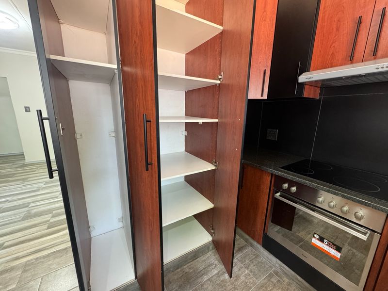 A modern kitchen featuring dark wood cabinetry, a built-in oven and hob, extractor fan, stone countertops, and ample pantry storage space.
