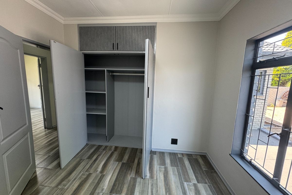 A bright, empty bedroom featuring modern wood-look tiled flooring, a built-in wardrobe with overhead storage, and a window fitted with security bars.