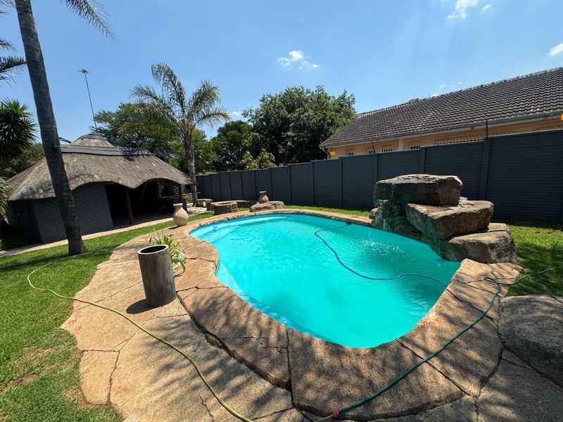 Outdoor pool area featuring a rock water feature, paved surround, and a thatched lapa, set within a private garden with perimeter walling.