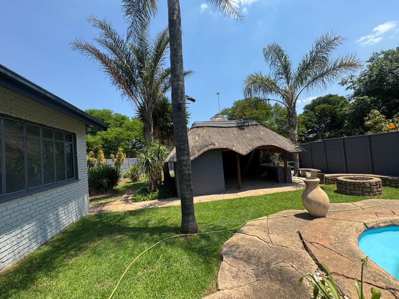 Outdoor entertainment area featuring a swimming pool, paved surround, built-in fire pit, and a thatched lapa structure set within a landscaped garden.