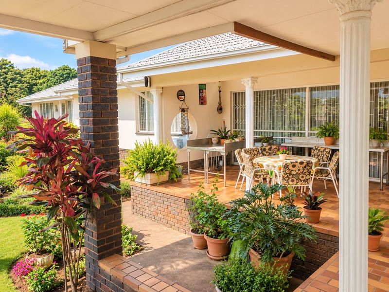 A spacious covered patio featuring brick columns, tiled flooring, and built-in prep counters, overlooking a well-maintained garden area.
