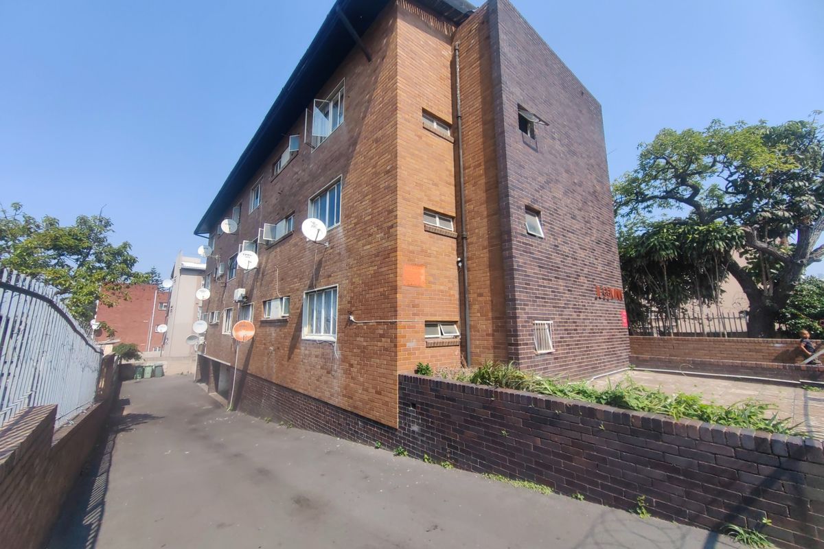Side and rear view of a multi-story brick apartment building featuring an asphalt driveway and perimeter brick boundary wall.