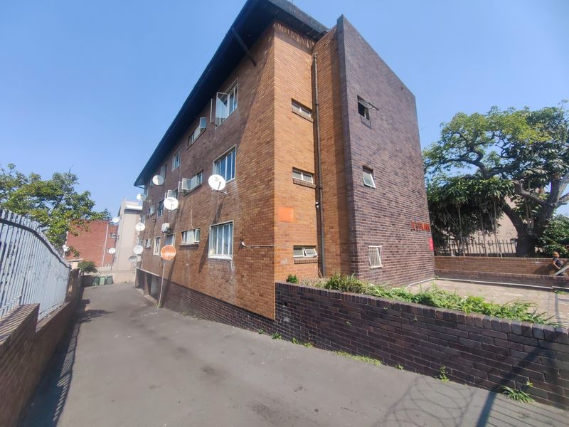 Side and rear view of a multi-story brick apartment building featuring an asphalt driveway and perimeter brick boundary wall.