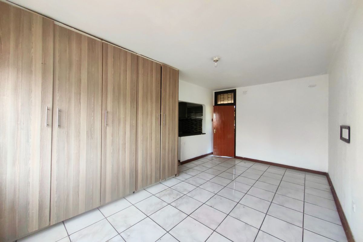 A spacious, empty bedroom featuring light wood-finish built-in cupboards, white tiled flooring, and a wooden entrance door.
