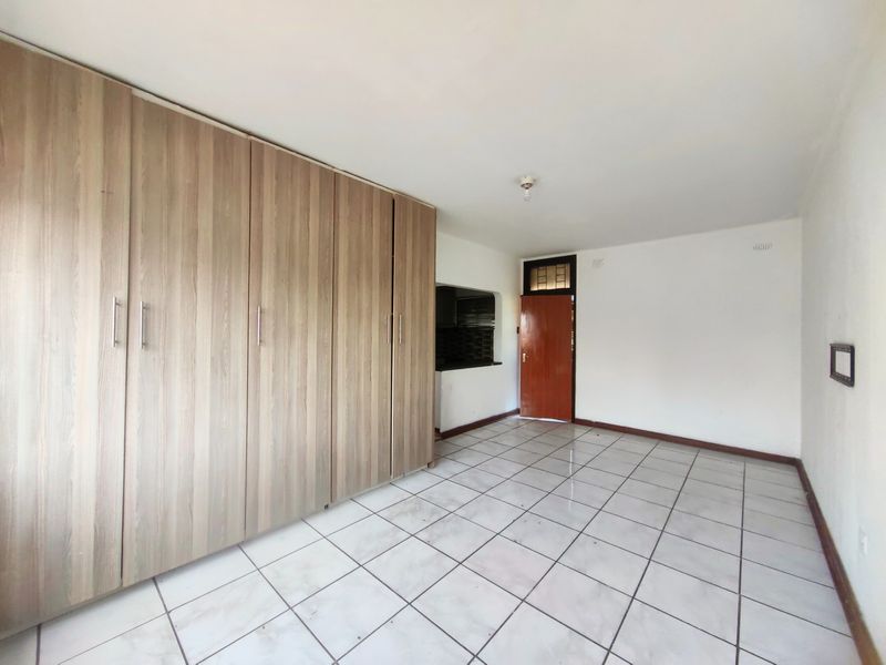 A spacious, empty bedroom featuring light wood-finish built-in cupboards, white tiled flooring, and a wooden entrance door.