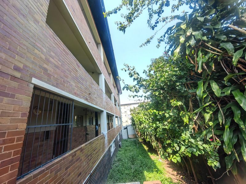 Rear view of a multi-story face brick residential building featuring external walkways, security bars, and an adjacent garden area with mature trees.