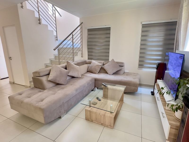 A bright and modern living room featuring tiled flooring, a staircase with a stainless steel balustrade, and dual roller blinds.