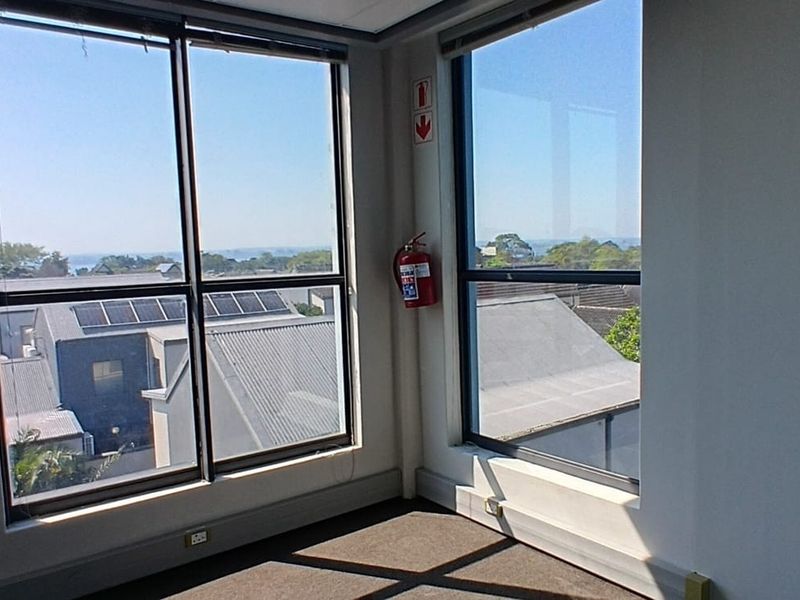 A bright, empty corner office space featuring large windows with natural light, carpeted flooring, and a wall-mounted fire extinguisher.