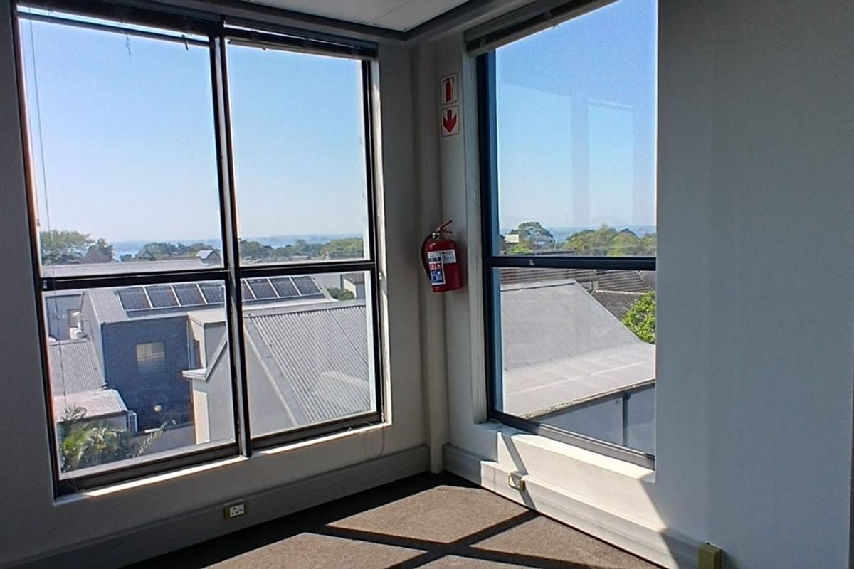 A bright, empty corner office space featuring large windows with natural light, carpeted flooring, and a wall-mounted fire extinguisher.