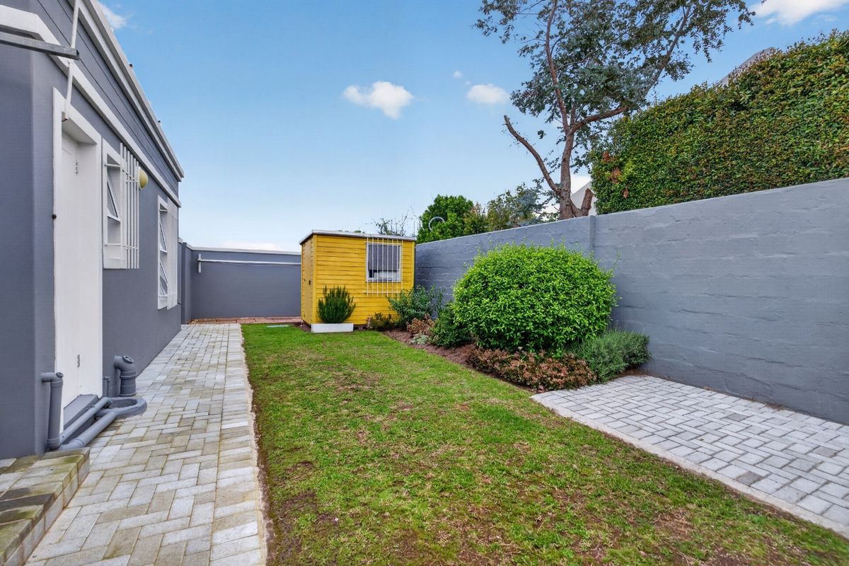 A neat rear garden featuring a paved walkway, a well-maintained lawn, and a yellow storage shed enclosed by a solid boundary wall.
