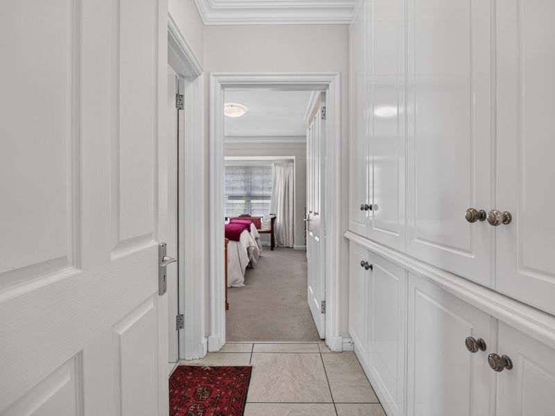 A bright, well-maintained hallway featuring extensive built-in white cabinetry, crown molding, and neutral tiled flooring, leading into an adjacent bedroom.