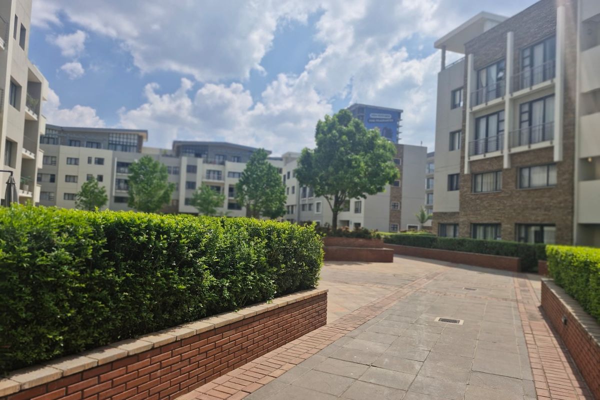 A well-maintained outdoor courtyard area featuring paved walkways, brick retaining walls, and landscaped hedges between multi-story apartment buildings.