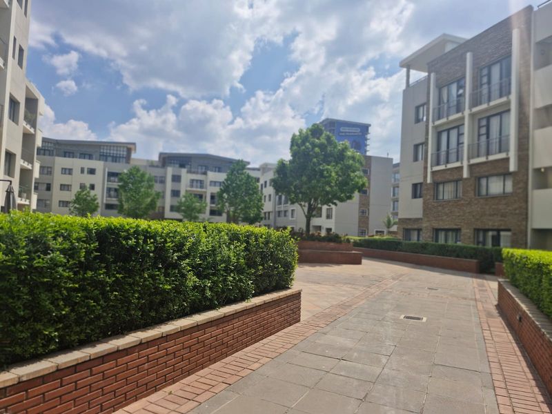 A well-maintained outdoor courtyard area featuring paved walkways, brick retaining walls, and landscaped hedges between multi-story apartment buildings.