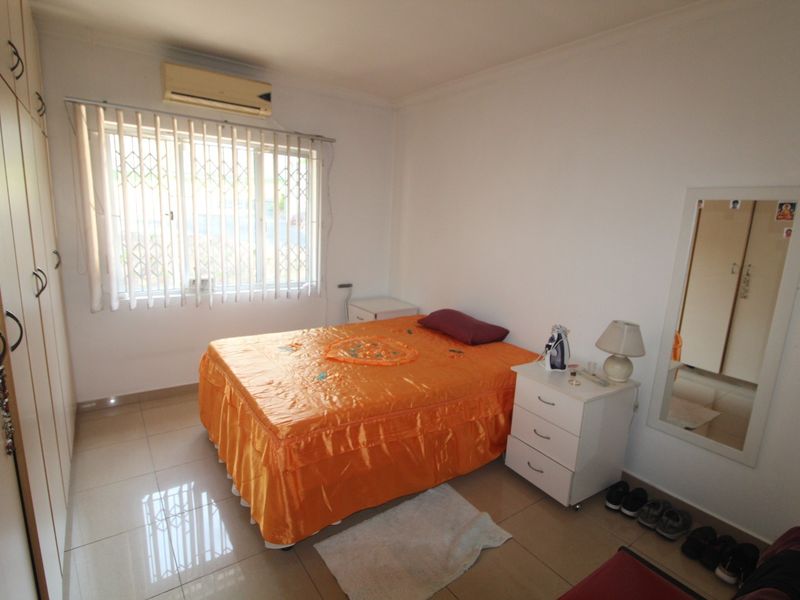 A functional bedroom featuring tiled flooring, built-in cupboards, and a wall-mounted air conditioning unit with security window treatments.