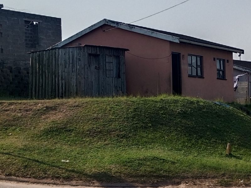 A single-story residential property featuring a painted exterior, corrugated metal roof, and an adjacent timber storage structure on a sloped grass lot.