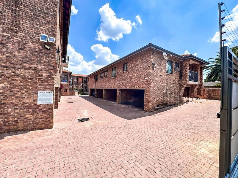 Exterior view of a face brick apartment complex featuring a paved driveway, covered parking bays, and secure perimeter fencing.