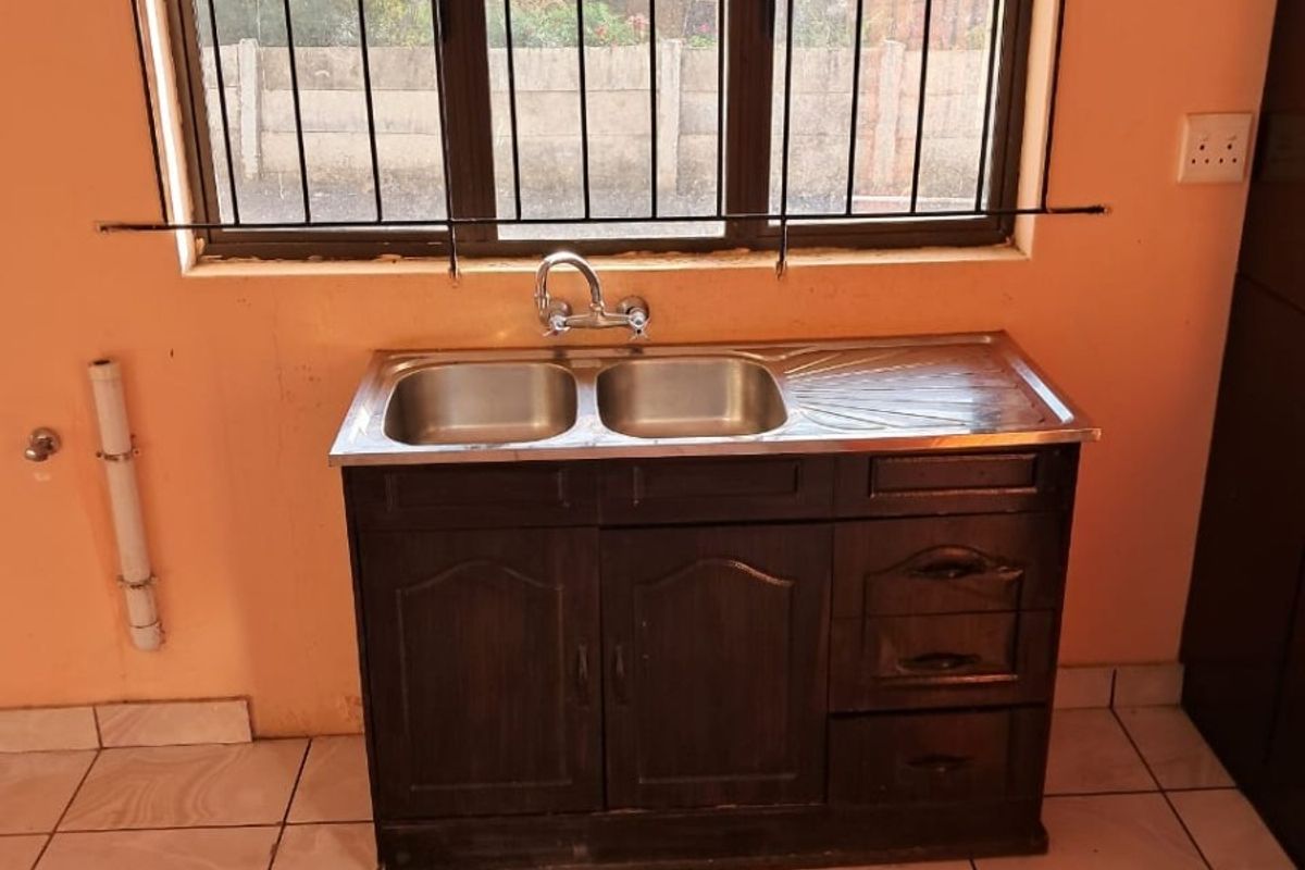 A basic kitchen area featuring a stainless steel double sink, dark wood cabinetry, and tiled flooring, situated beneath a window with security bars.