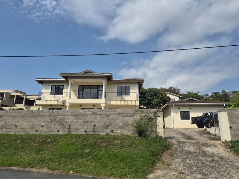 Front view of a two-story house featuring a balcony, tiled roof, and a detached garage with a concrete driveway and block boundary wall.