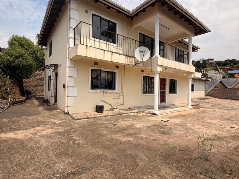 Rear view of a two-story house featuring a balcony with wrought iron railings, concrete columns, and a large concrete driveway area.