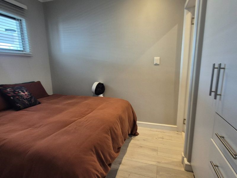 A bedroom featuring light-toned laminate flooring, built-in cupboards with modern handles, and a window fitted with venetian blinds.