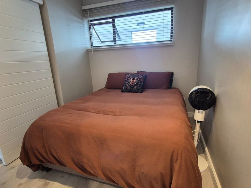 A compact bedroom featuring light-toned laminate flooring, a sliding door, and a window fitted with venetian blinds.