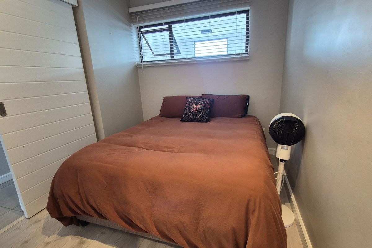 A compact bedroom featuring light-toned laminate flooring, a sliding door, and a window fitted with venetian blinds.