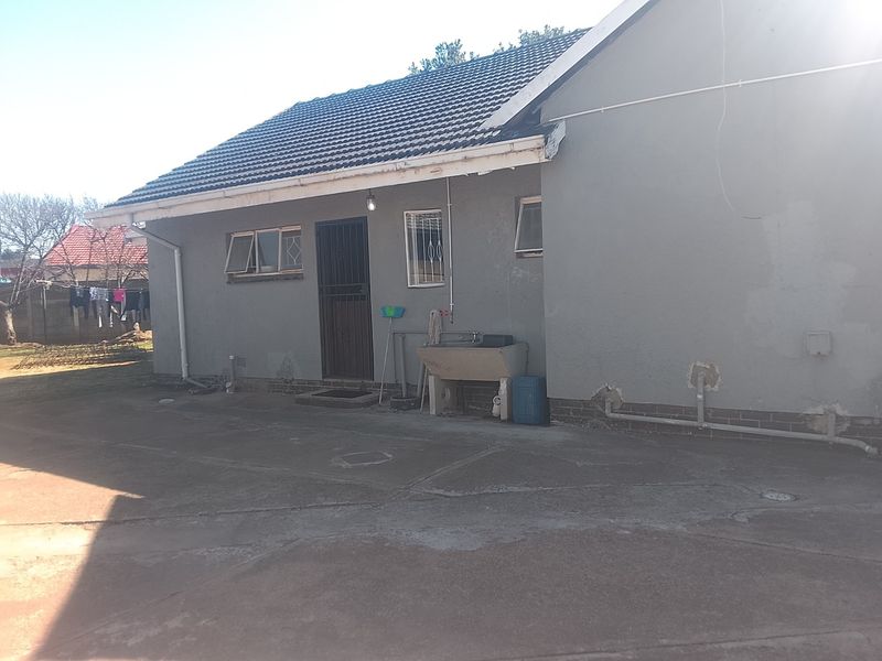 Rear exterior view of the house featuring a concrete apron, external laundry sink, and security gate, showing signs of wear to the plaster and guttering.