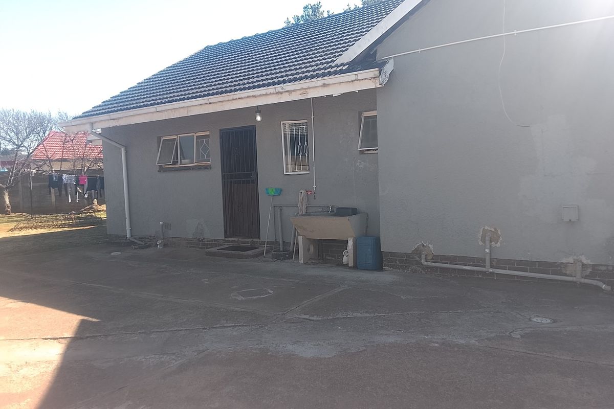 Rear exterior view of the house featuring a concrete apron, external laundry sink, and security gate, showing signs of wear to the plaster and guttering.