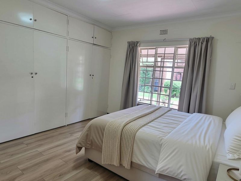 A well-maintained bedroom featuring light laminate flooring, extensive built-in cupboard storage, and a window with security bars.
