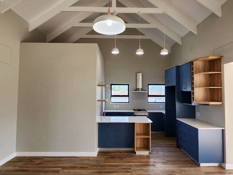 Modern open-plan kitchen and living area featuring a high vaulted ceiling with exposed trusses, contemporary blue cabinetry, and durable laminate flooring.
